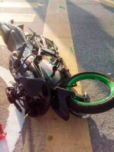 Photograph 4: General view of The Motorcycle at the scene of collision taken by passers-by. Note the rim of the broken front wheel that was slightly bent towards the nearside (red arrow), relatively more severe damage on the offside aspect of the front fairing (yellow arrow), broken nearside front signal lamp (light blue arrow) and the faint scratch mark on the surface of the yellow box (green arrows) indicating that The Motorcycle was dragged from its initial position closer to the middle of the North-bound lanes of Jalan Semarak at the South side of The Junction, near the position of the third yellow bar of the zebra crossing (white arrow) to the West of the raised median.